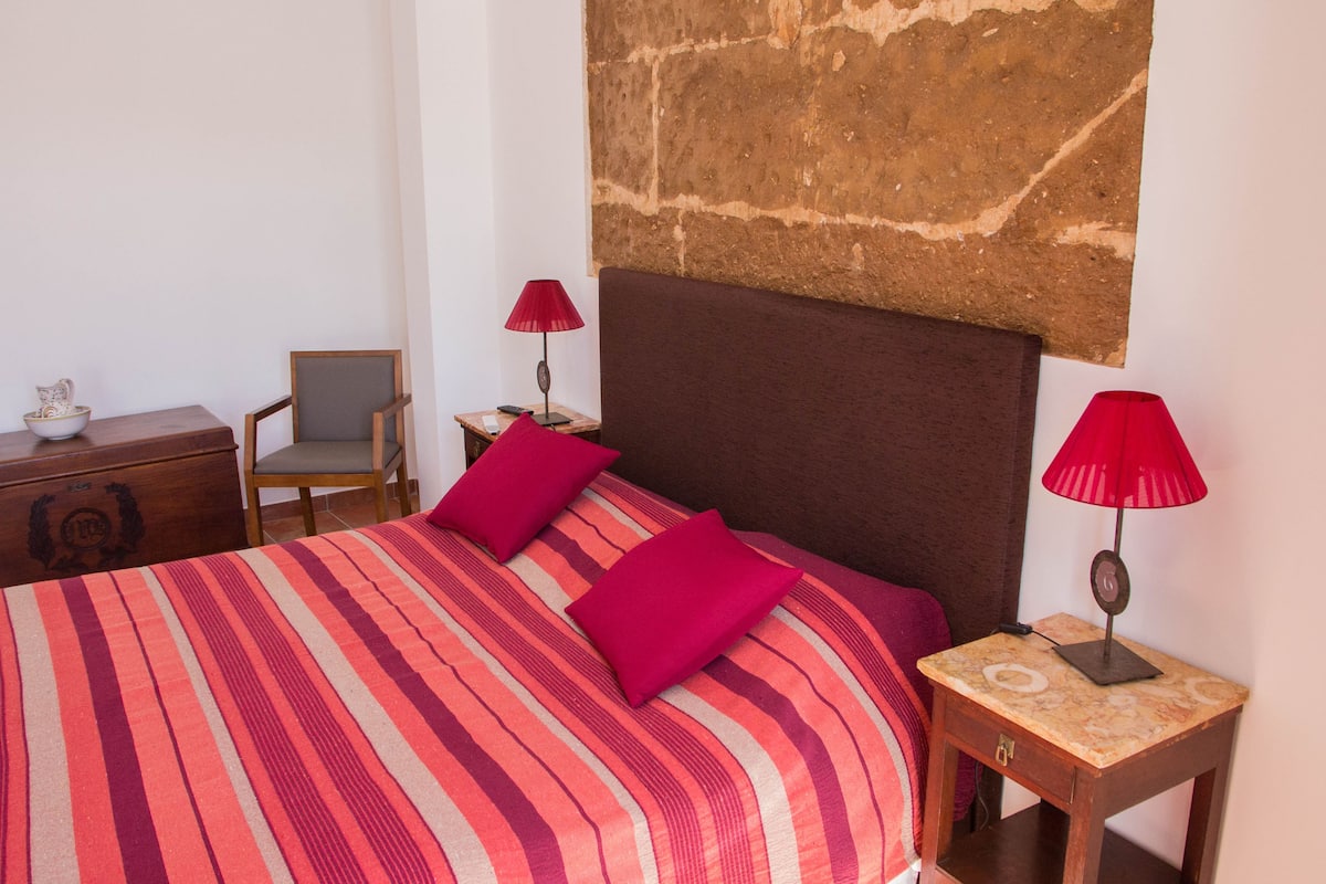 A bed with a striped bedspread and two red pillows, a wooden nightstand with a lamp, a chair, and a small table with a bowl and a book.