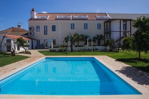 A rectangular swimming pool with a tiled deck, surrounded by a well-maintained lawn and a building with a tiled roof and multiple windows. - Casas da Moagem Turismo Rural (Santiago do Cacém)