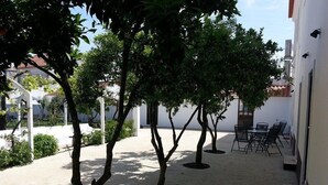 Un espace extérieur aménagé en patio, avec un mur blanc, une table entourée de chaises et plusieurs arbres.