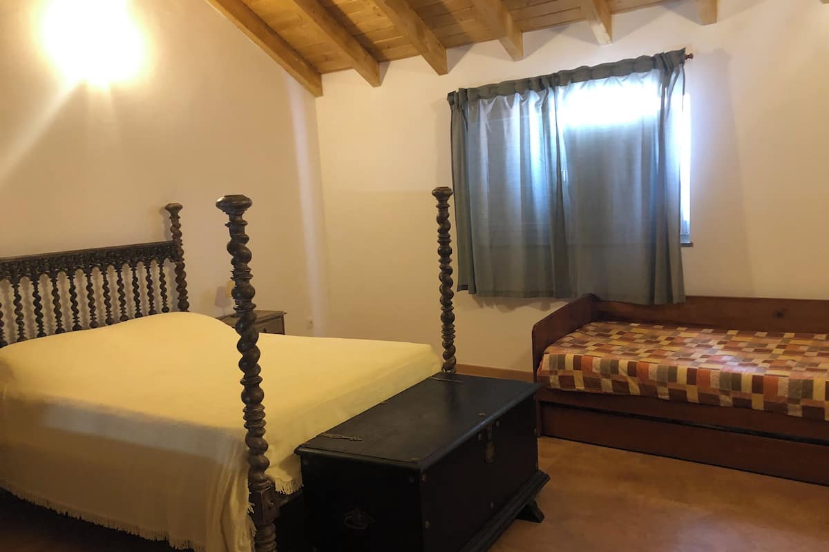 A bedroom with a four-poster bed, a wooden bench, a small table, a window with curtains, and a checkered bedspread.