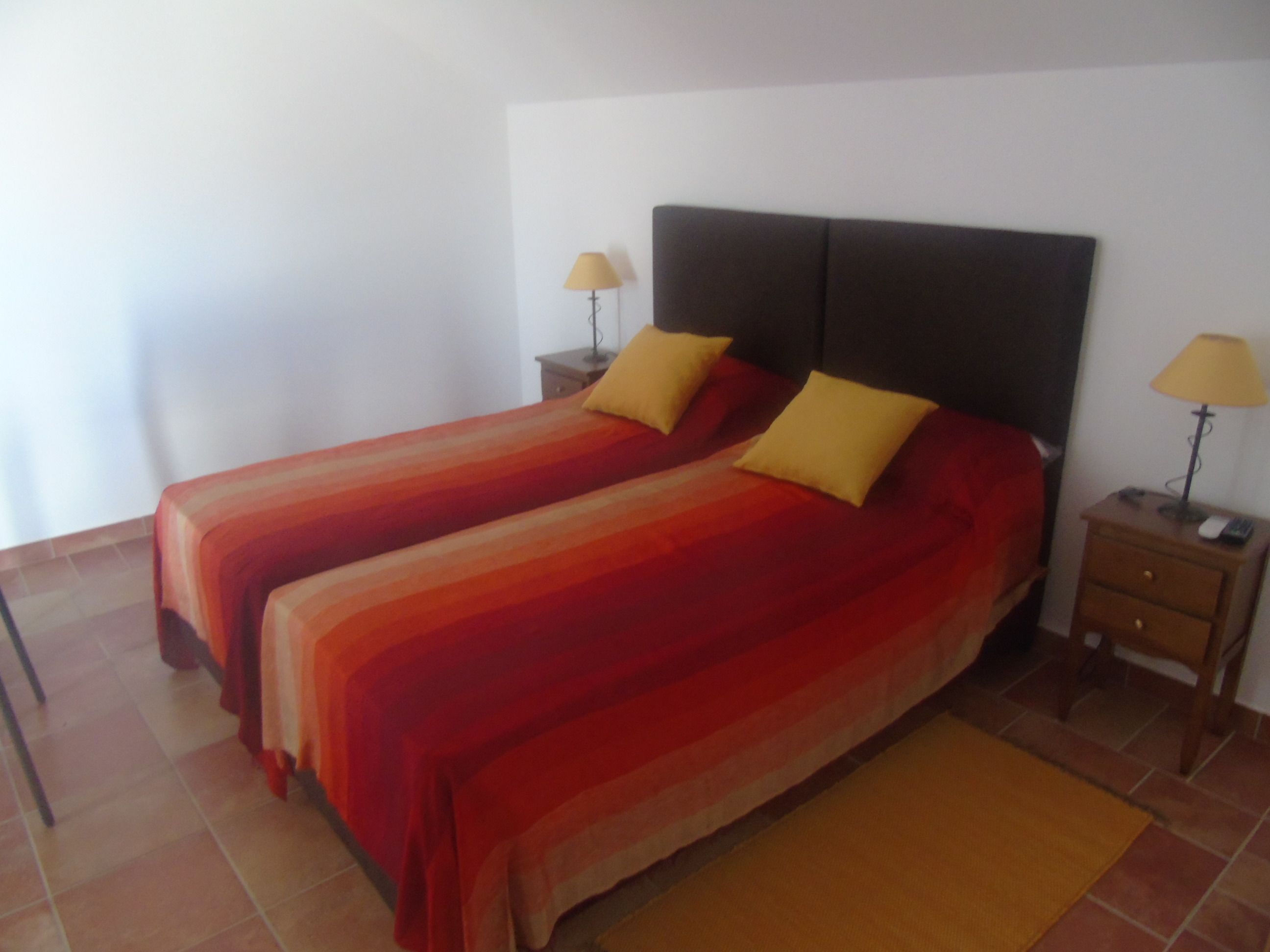 A double bed with a striped bedspread and two pillows, a nightstand with a lamp, and a small table with a lamp.