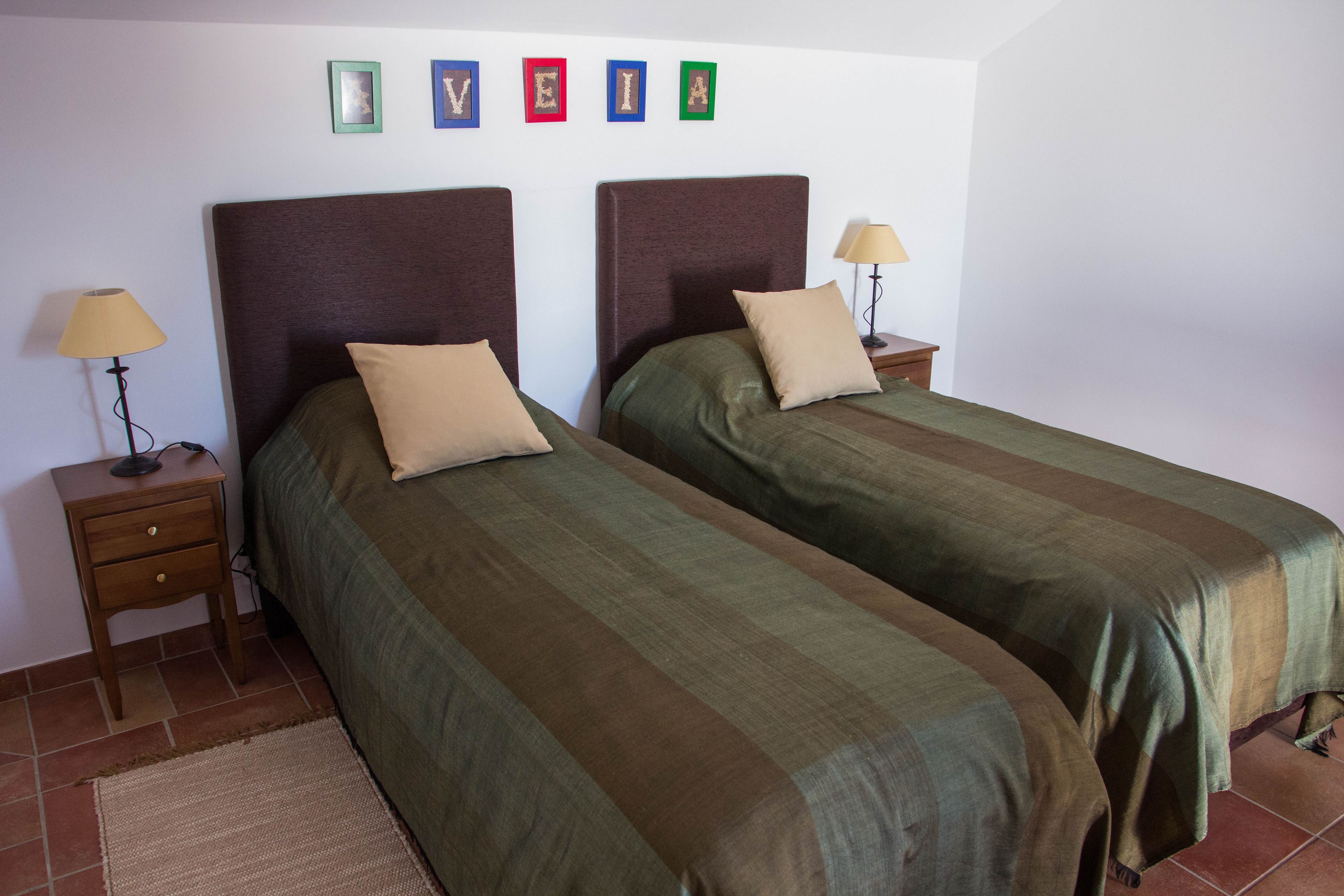 Quarto com duas camas, cada uma com cabeceira, mesa de cabeceira e luminária, além de um tapete no chão.