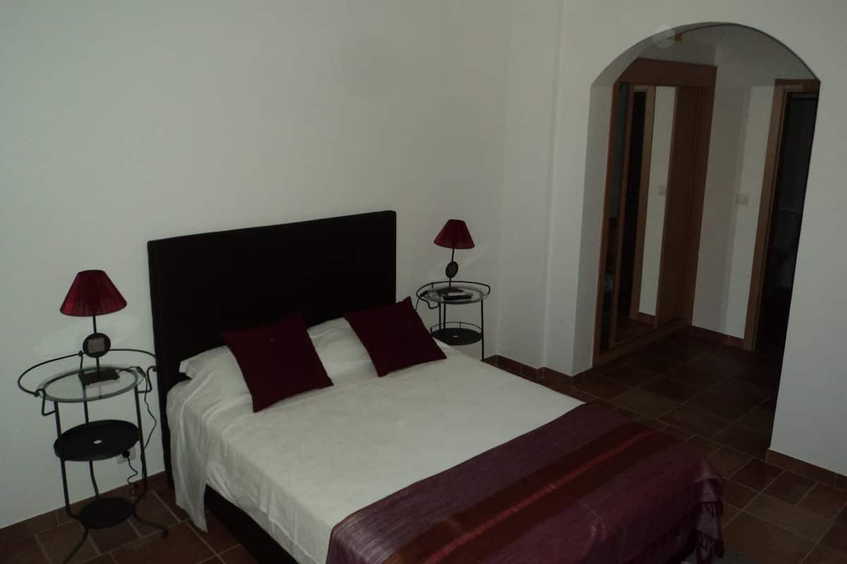 A bedroom with a bed, two bedside tables with lamps, a red throw blanket, and a door leading to another room.