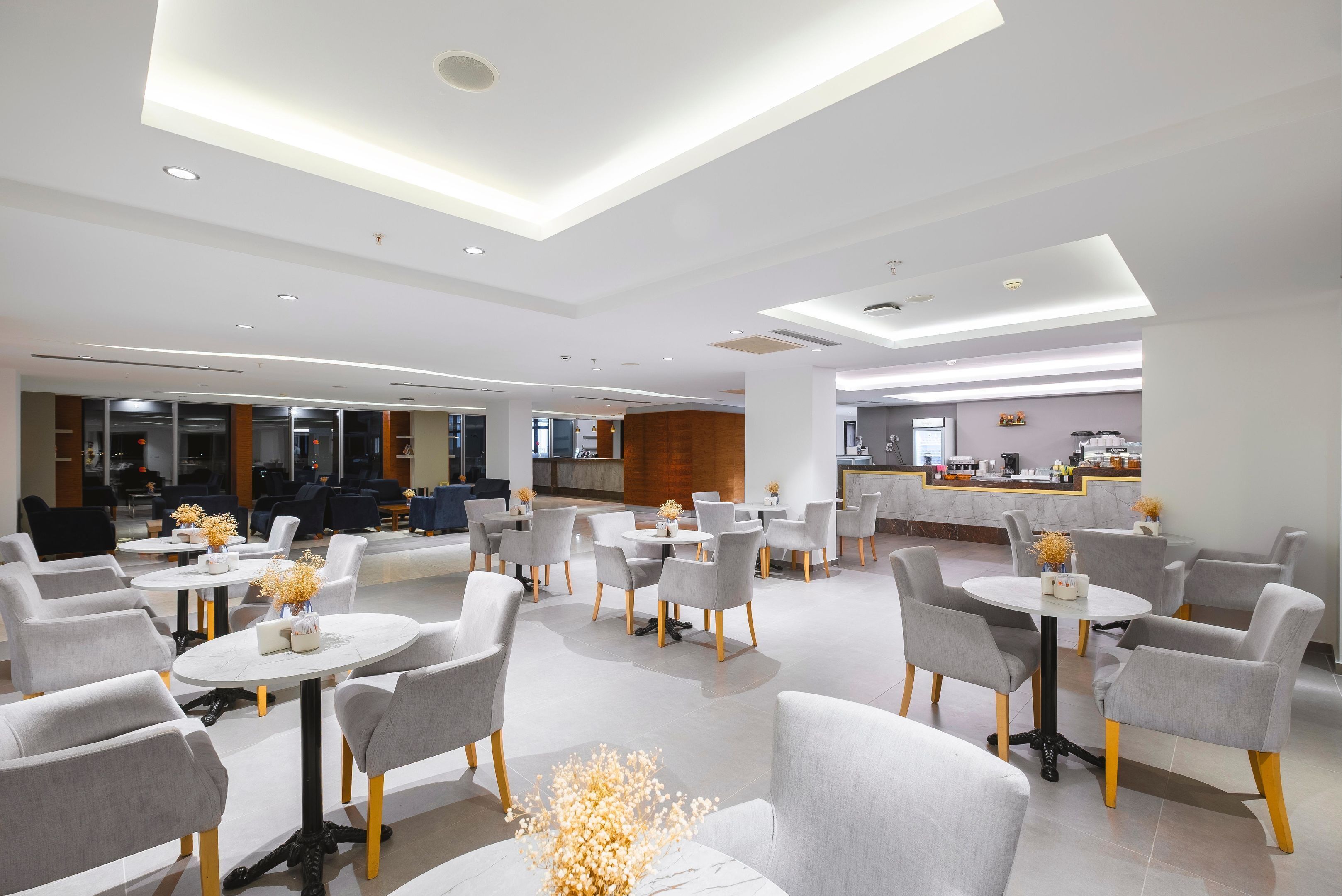 A modern hotel lobby with round tables and grey chairs, a reception desk, and a kitchen area in the background.