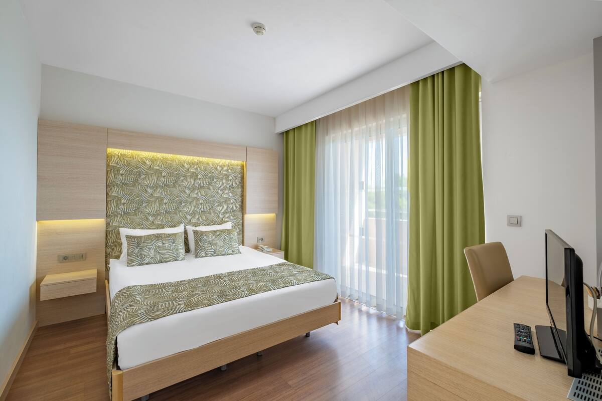 A modern hotel room with a large bed, a desk with a computer, and a view of the outdoors.