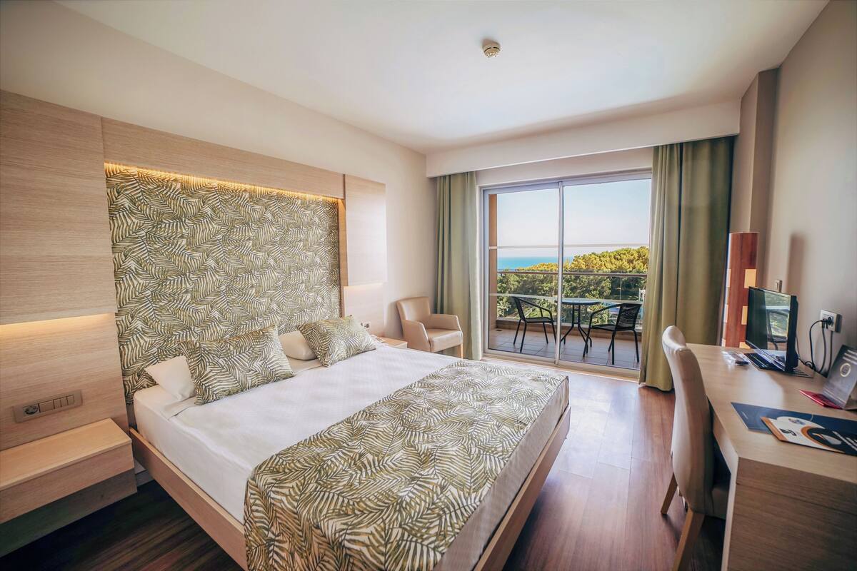 A modern hotel room with a large bed, a desk, and a balcony with a view.