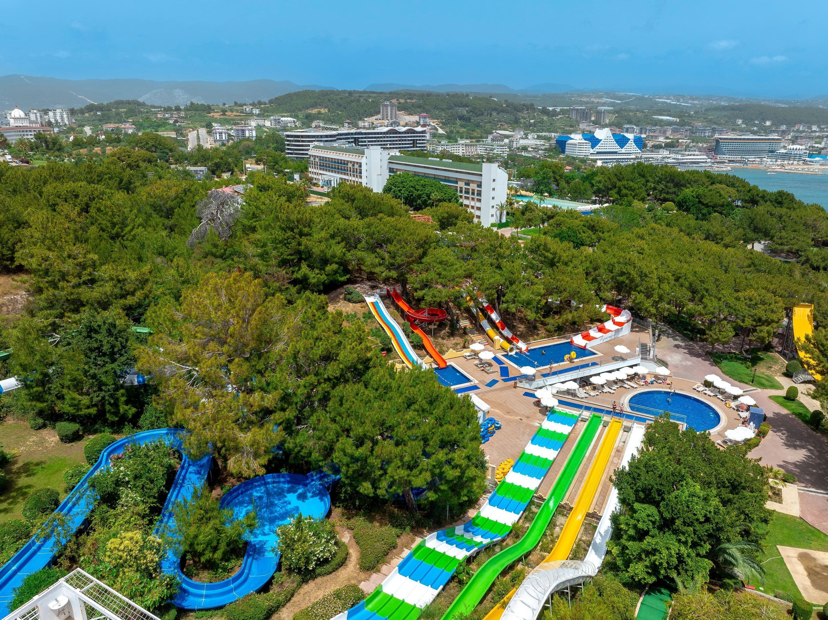 Parque acuático con múltiples toboganes y piscinas, rodeado de follaje y edificios al fondo.