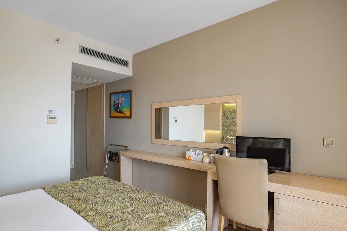 A hotel room with a bed, a desk with a TV, a chair, a coat rack, and a painting on the wall.