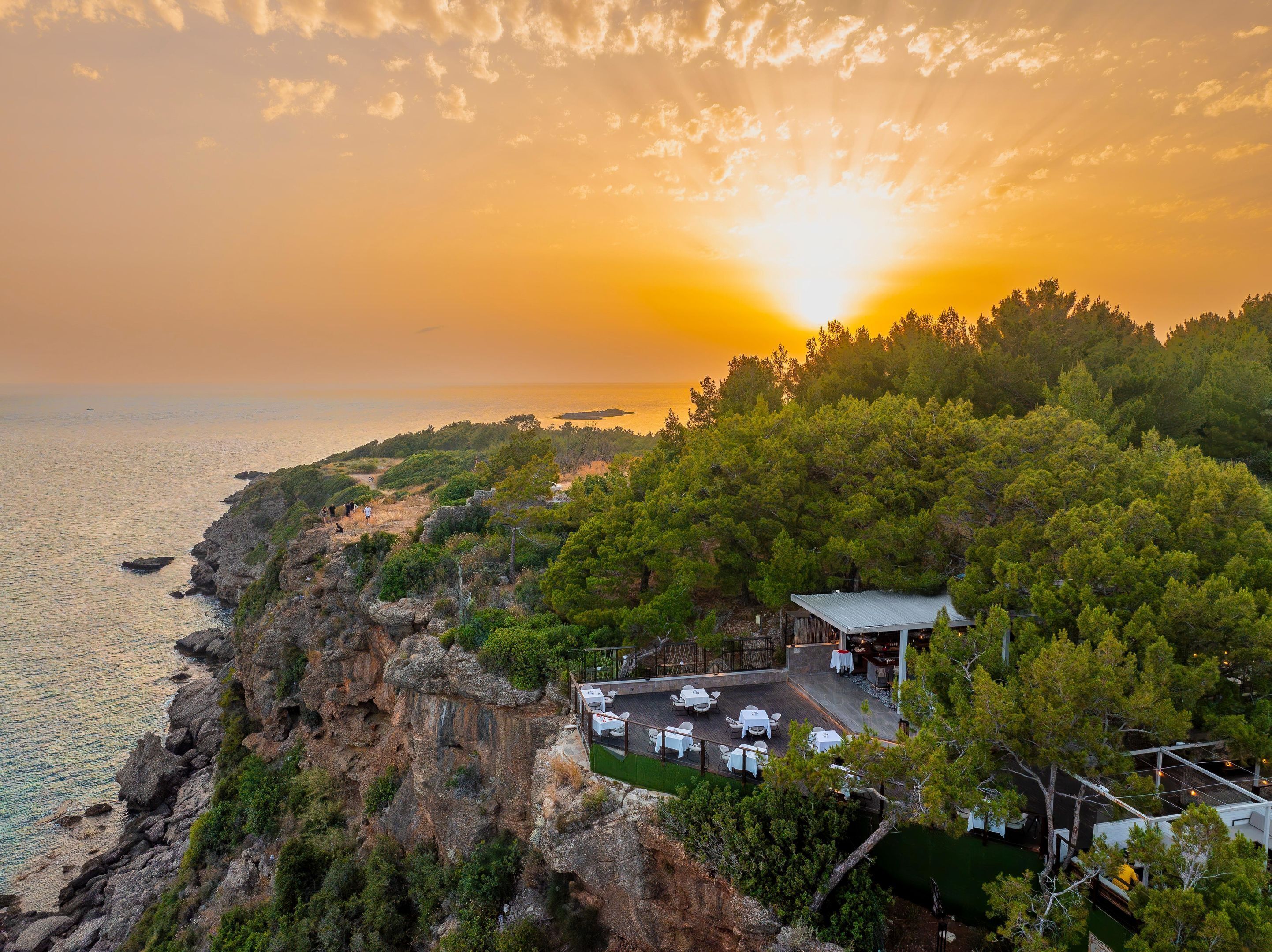 A coastal property with a dining area overlooking the sea, surrounded by dense greenery and rocky cliffs.