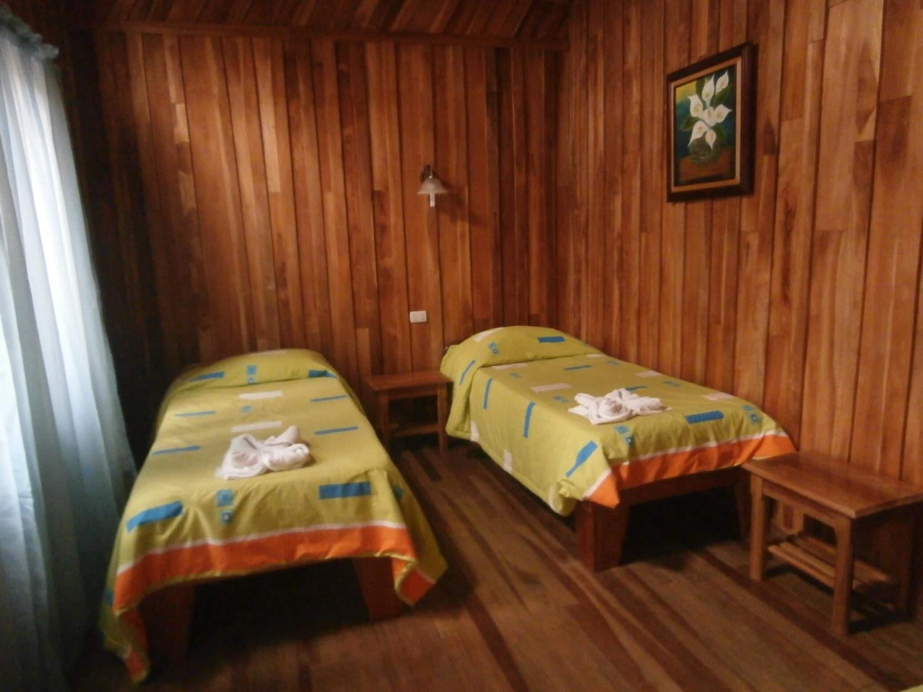 A room with two single beds, wooden walls, a small table, and a framed picture on the wall.