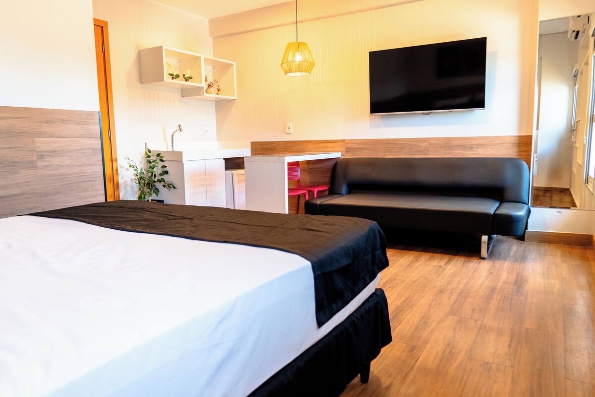 Una habitación de hotel moderna con una cama grande, un sofá, un televisor montado en la pared y una pequeña kitchenette.