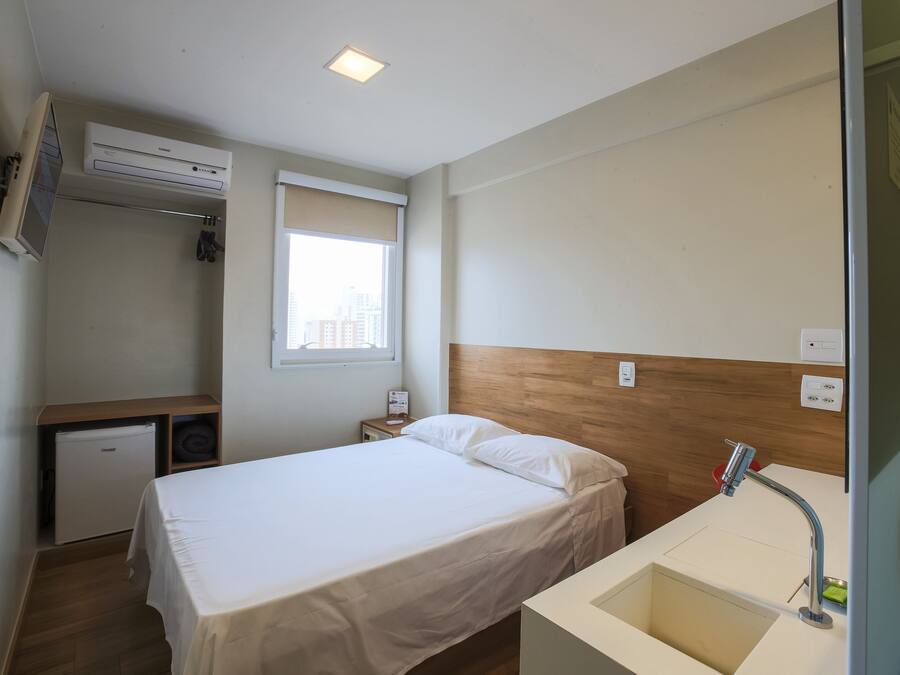 Una habitación pequeña y limpia con cama, escritorio, mini-refrigerador y ventana con vista a la ciudad.