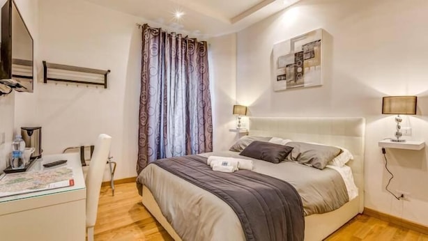 Une chambre d’hôtel avec un lit, des lampes de chevet, un bureau, une chaise, un miroir et une fenêtre avec des rideaux.