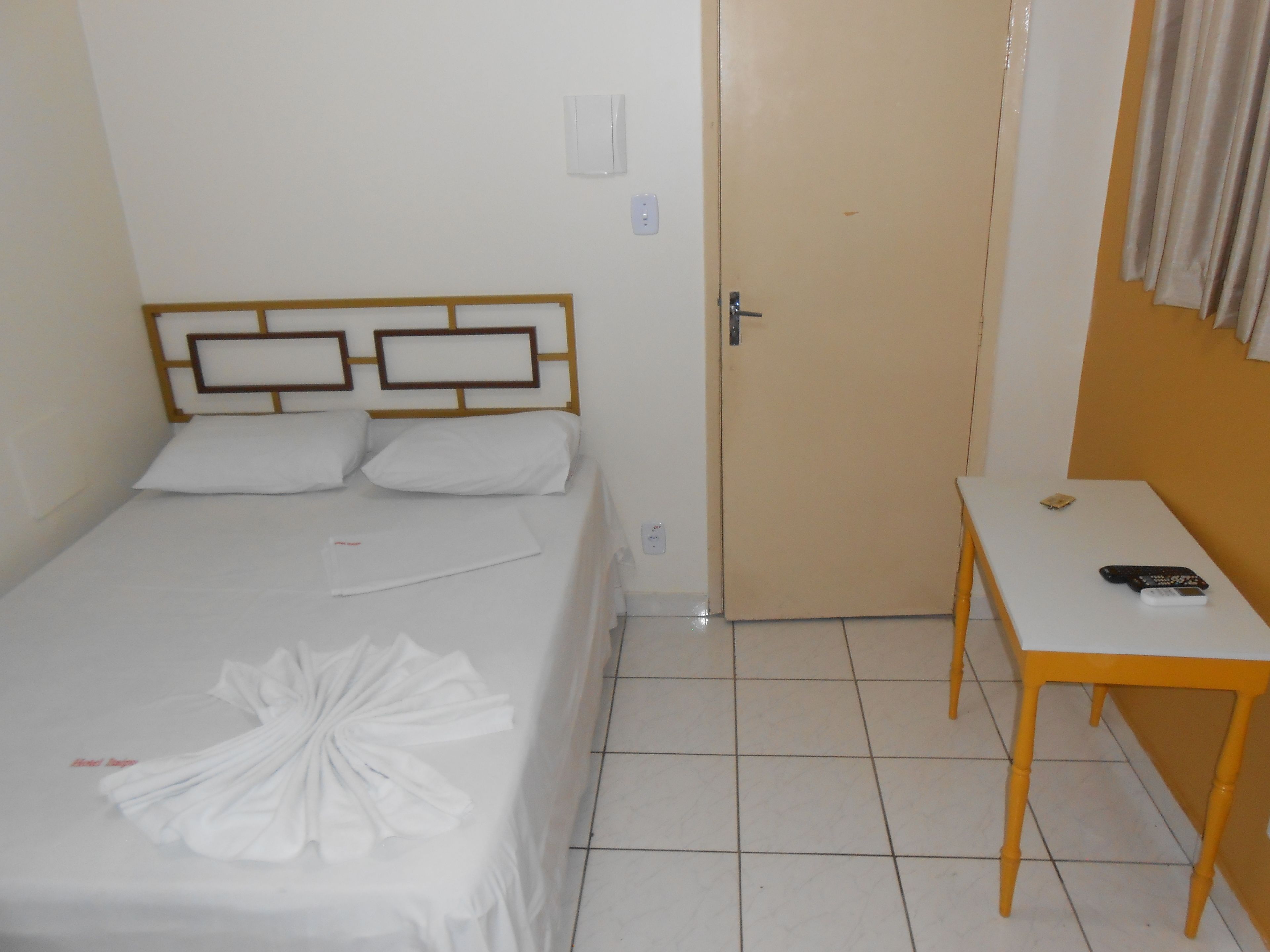 Uma cama de solteiro com cabeceira de madeira, duas almofadas e uma toalha branca dobrada sobre a cama. Há uma pequena mesa com um controle remoto.
