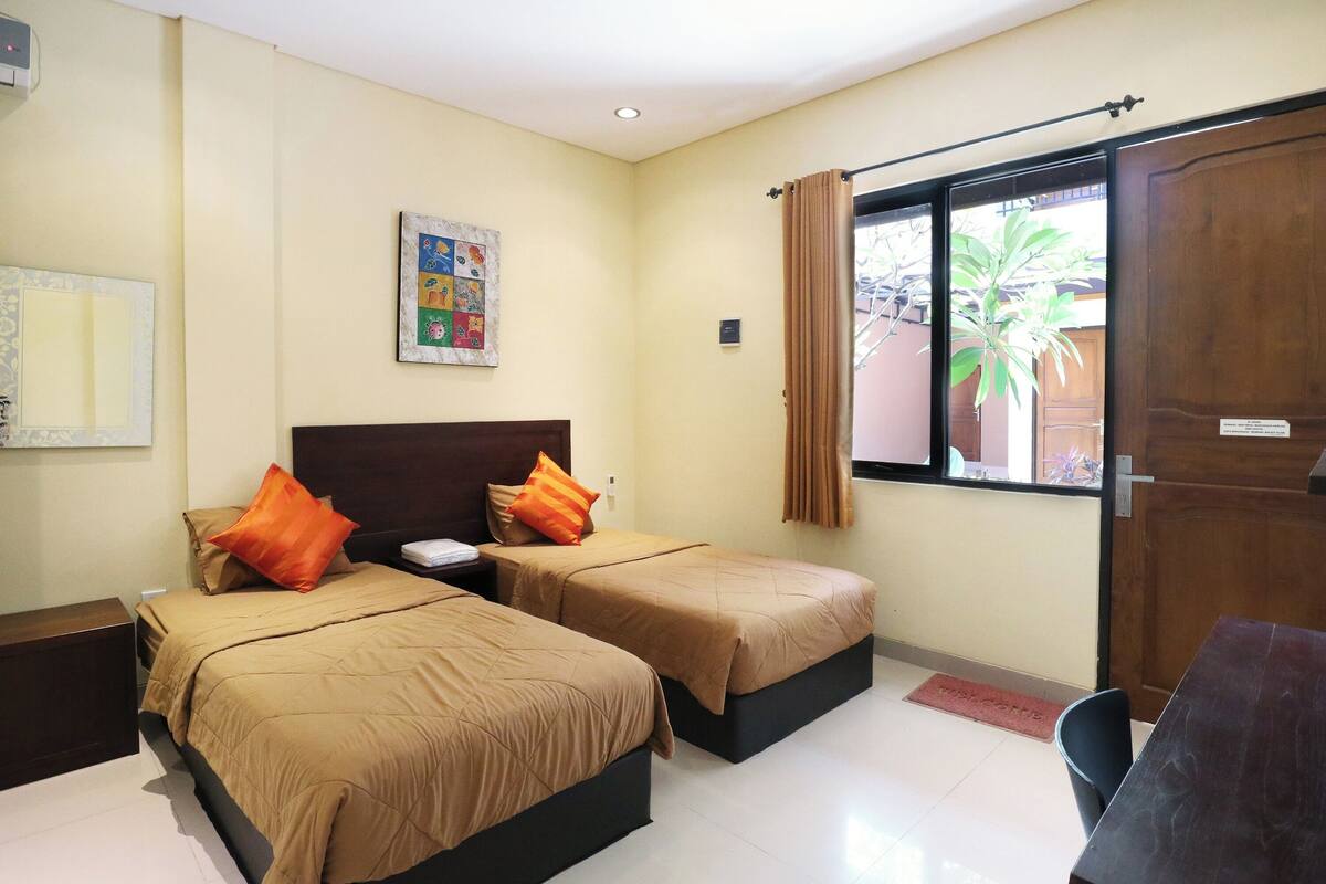 A hotel room with two beds, a wooden door, a window with curtains, a small table, and a wall-mounted artwork.
