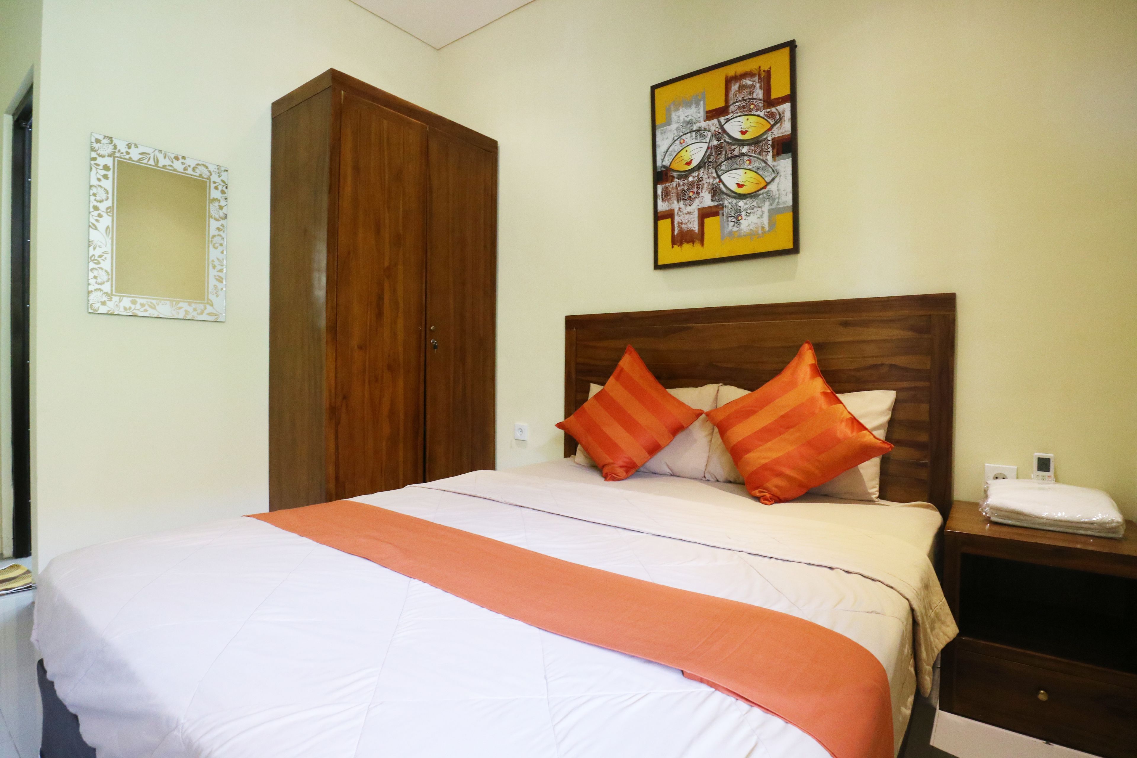 A neatly made bed with a white and orange bedspread, two orange pillows, a wooden wardrobe, and a framed artwork on the wall.