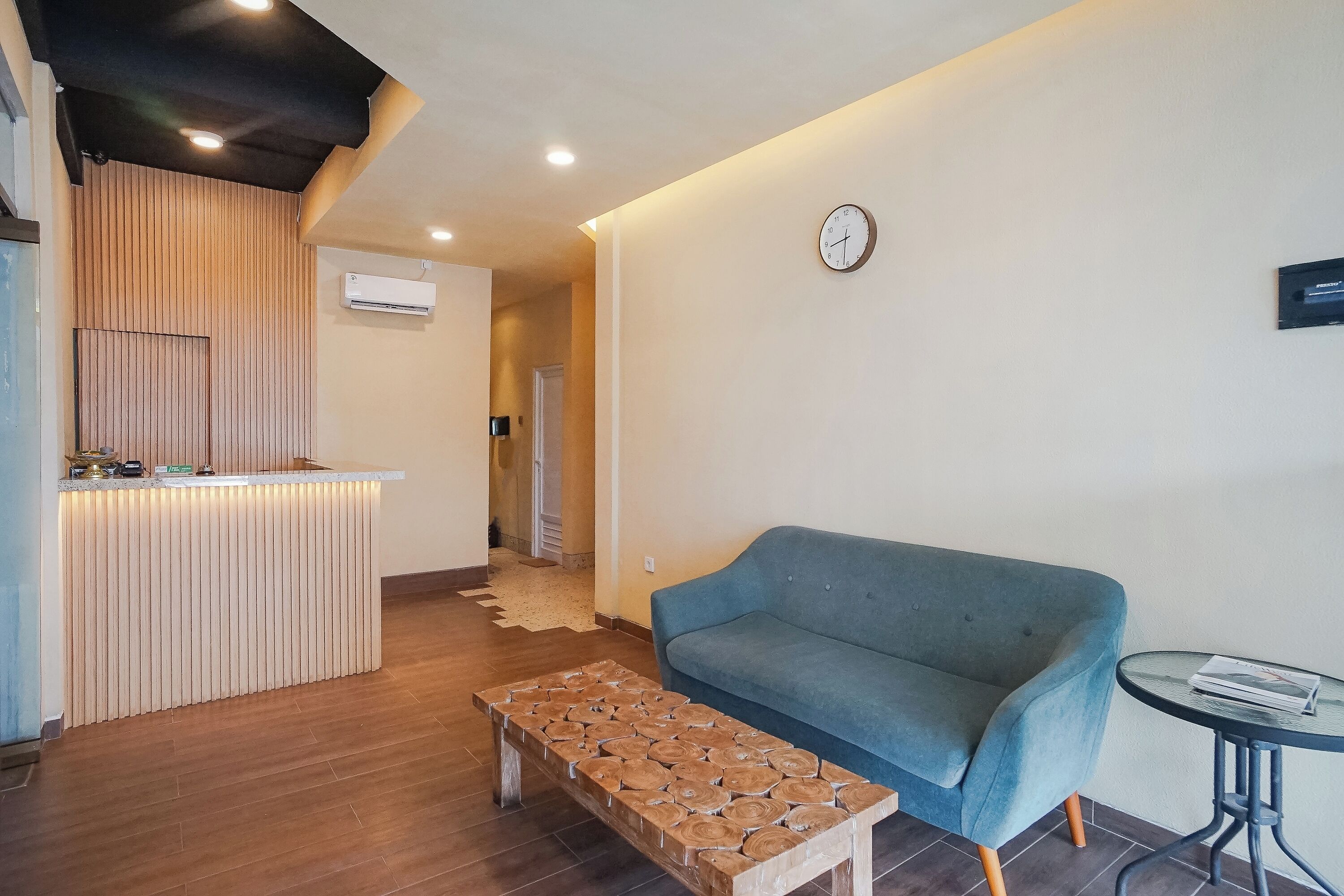 A modern living area with a blue sofa, a wooden coffee table, a small round table with a book, a wall clock, and an air conditioning unit.