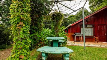 A rustic outdoor area with a green table and bench, surrounded by lush greenery and a red building with a window and door.