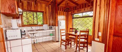 A wooden cabin with a kitchen area featuring a refrigerator, a microwave, and a sink. There is a dining table with chairs and a window with a view of greenery outside.