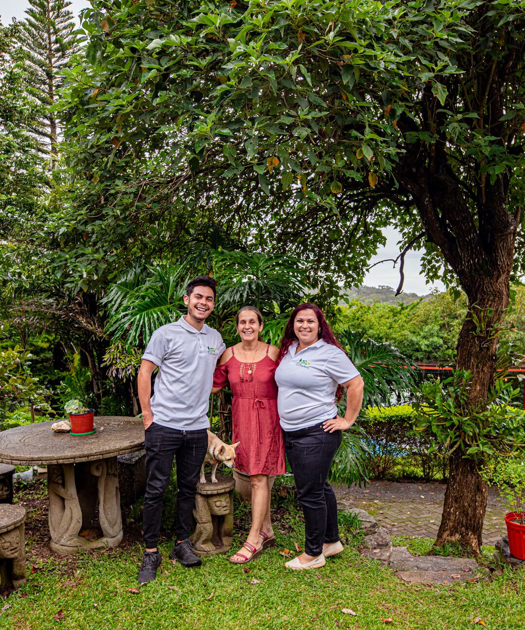Drie personen staan in een tuin met een stenen tafel en bloempotten.