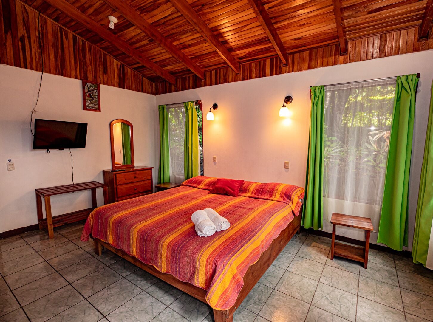 A bedroom with a wooden ceiling, a bed with a colorful striped bedspread, a wooden nightstand, a television mounted on the wall, and a window with green curtains.