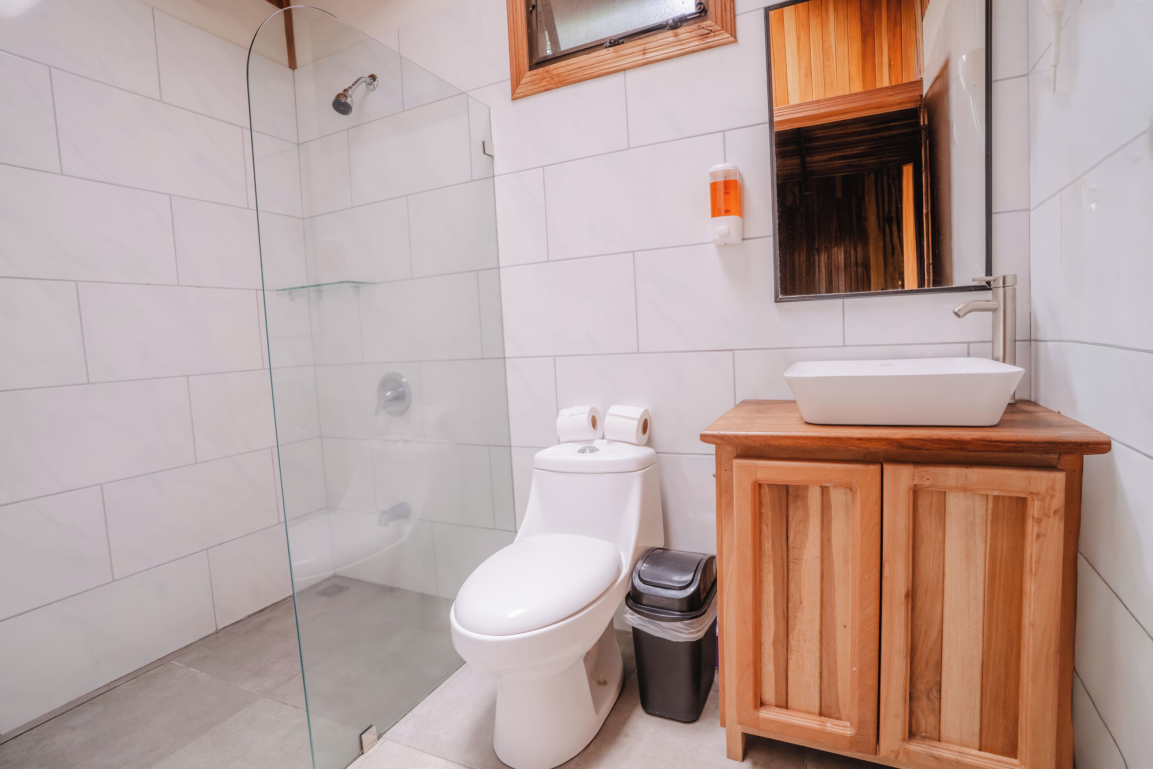 Een badkamer met een glazen douchecabine, een wit toilet, een houten wastafelmeubel met een wastafel en een prullenbak.