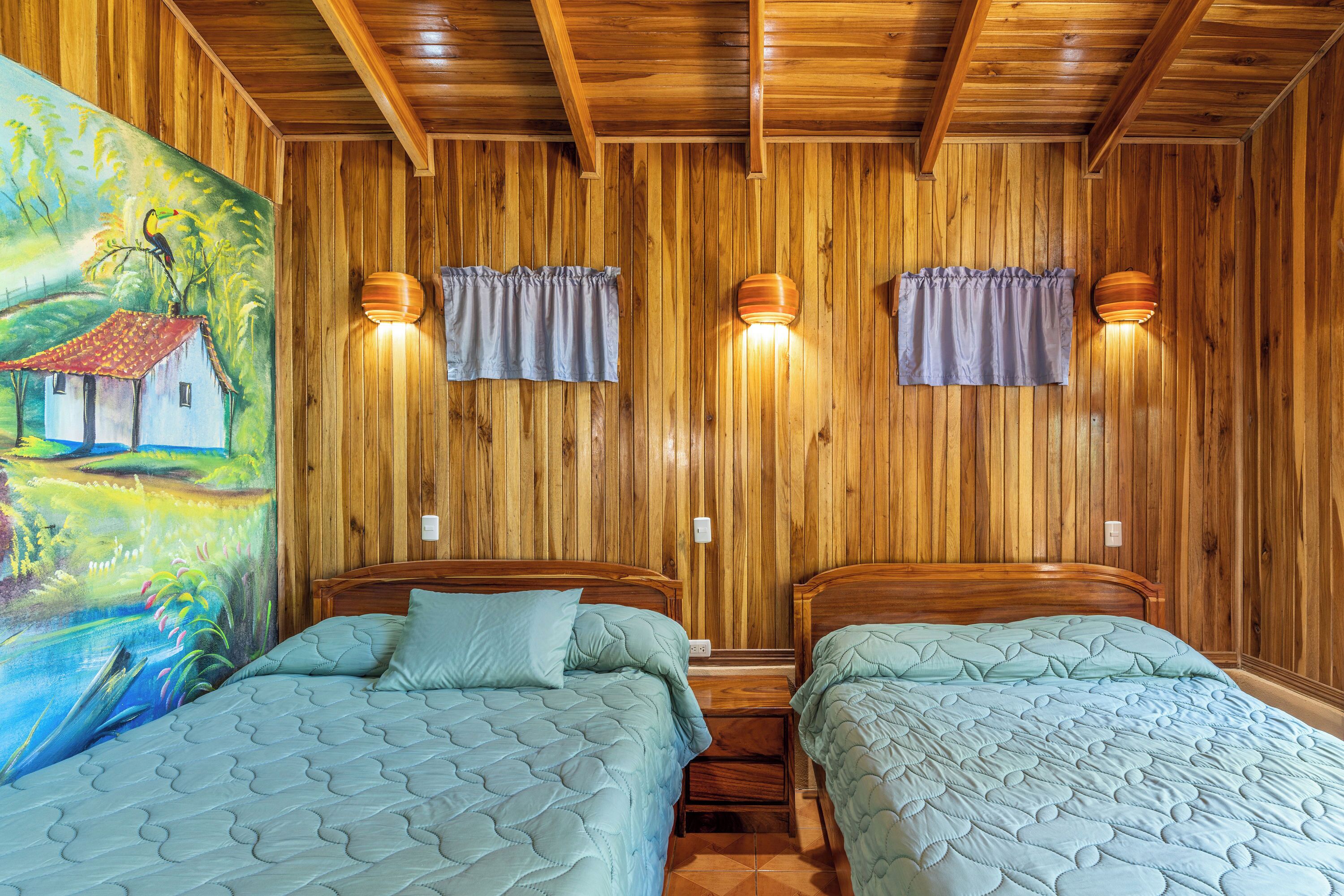 Une pièce aux murs lambrissés, avec deux lits, chacun recouvert d’une courtepointe matelassée et agrémenté d’un oreiller. Deux lampes sont fixées au mur et un tableau orne le mur de gauche.