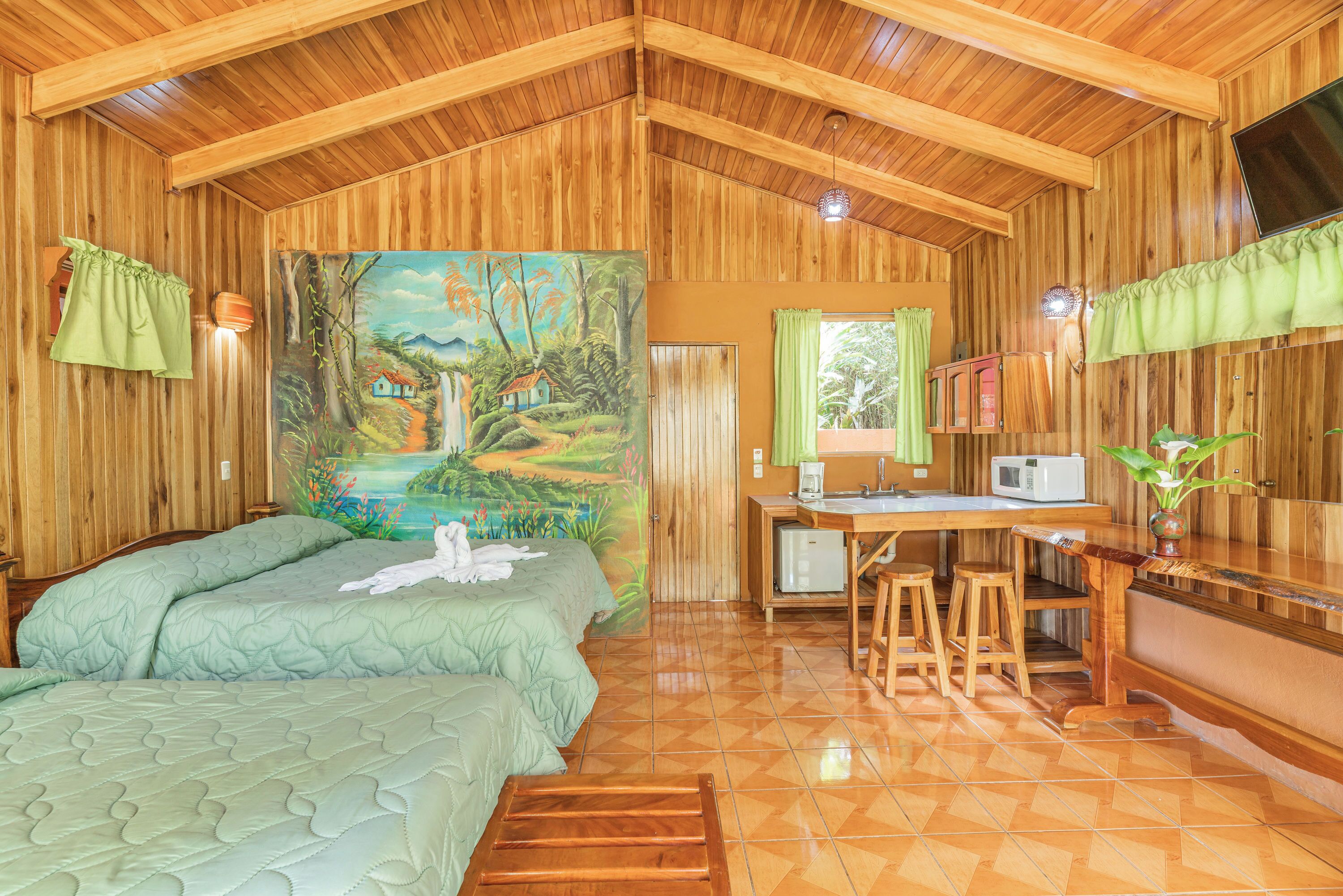 Una cabaña de madera con dos camas, un cuadro de una escena tropical, un rincón de cocina y una zona de comedor.