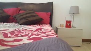 Una cama bien hecha con una colcha estampada, un cabecero y almohadas. Hay una mesita de noche con una lámpara y un cubo rojo.