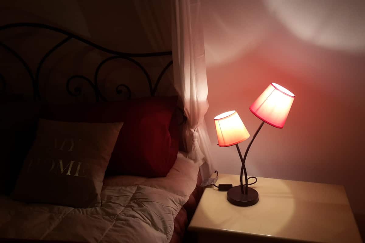 A bed with a white comforter and a pillow with "TOM" on it. There is a nightstand with a lamp and a small table beside it.