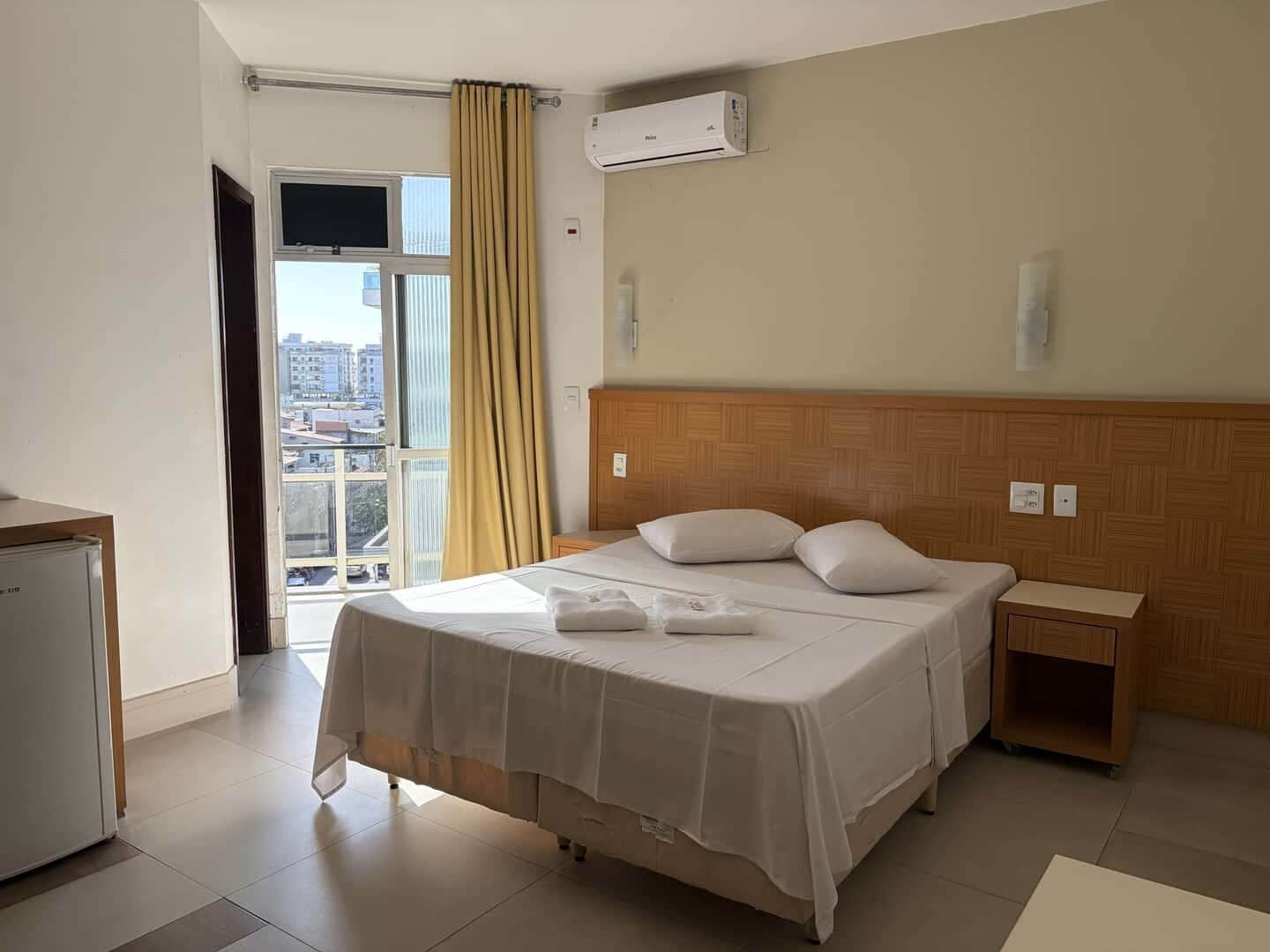 Habitación de hotel con cama, mesita de noche, aire acondicionado de pared y ventana con cortinas.