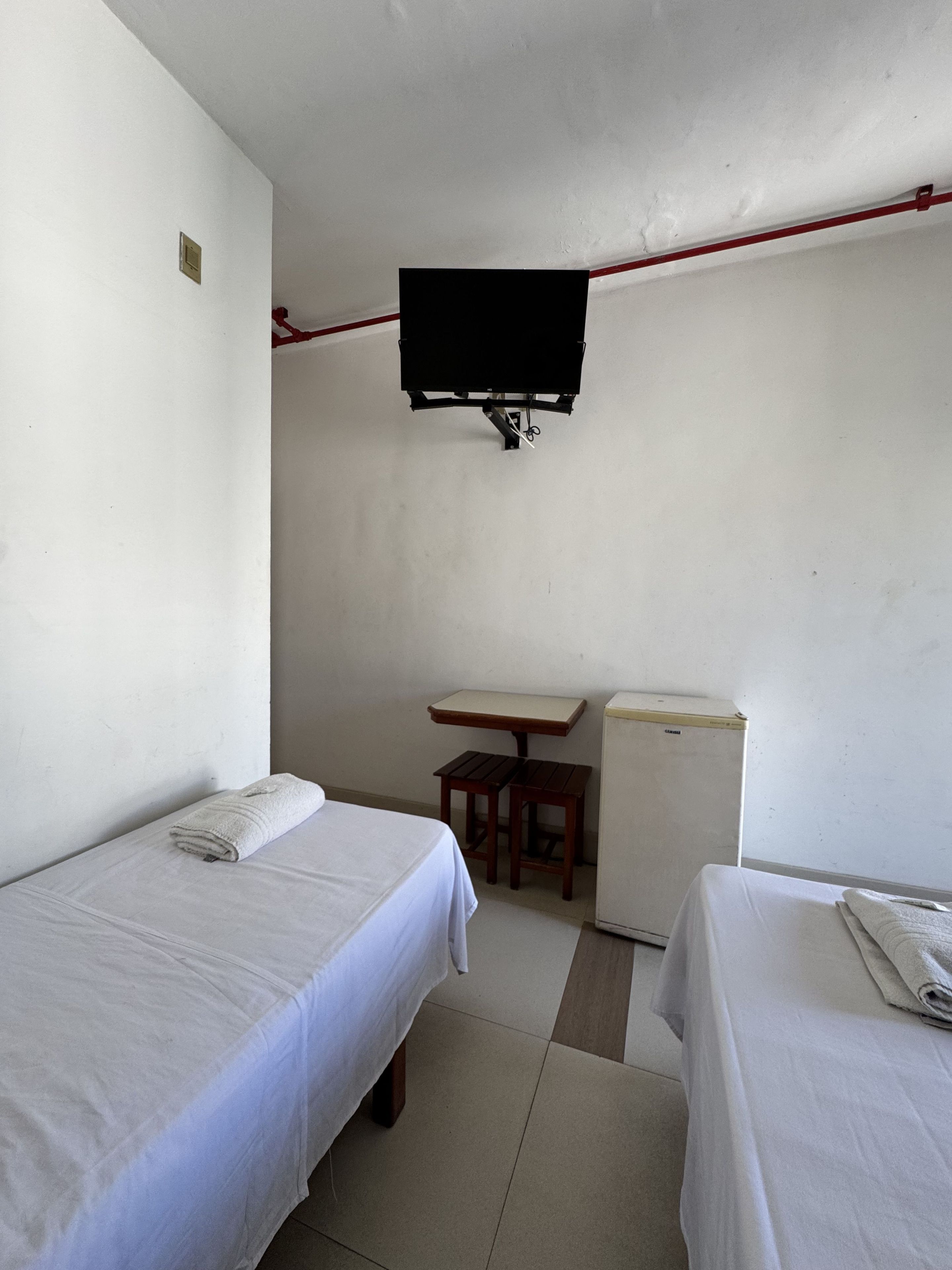A small, sparsely furnished room with two beds, a small table, and a television mounted on the wall.