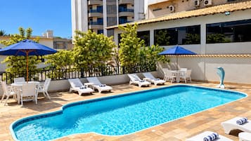 A swimming pool with lounge chairs and umbrellas, surrounded by a tiled deck and a low fence.