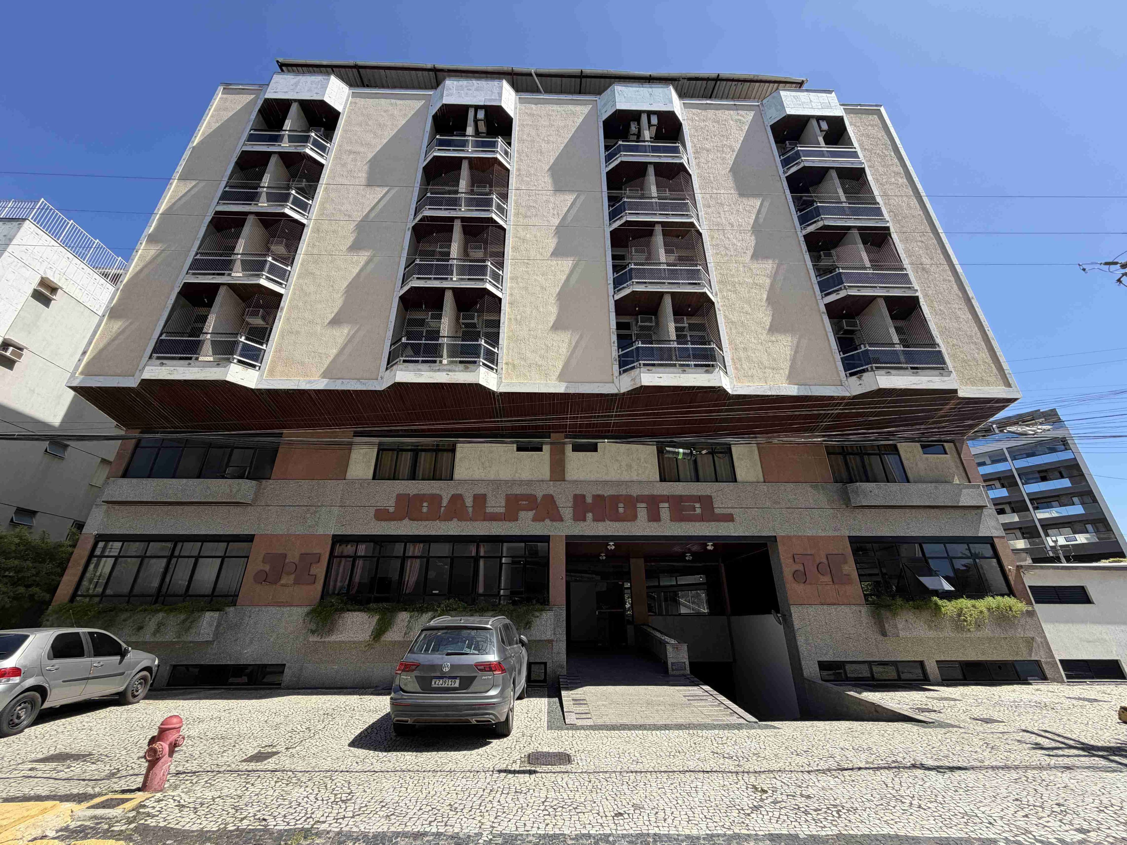 Un edificio de varios pisos con un llamativo letrero que dice 'JOALPA HOTEL', un estacionamiento y un hidrante.