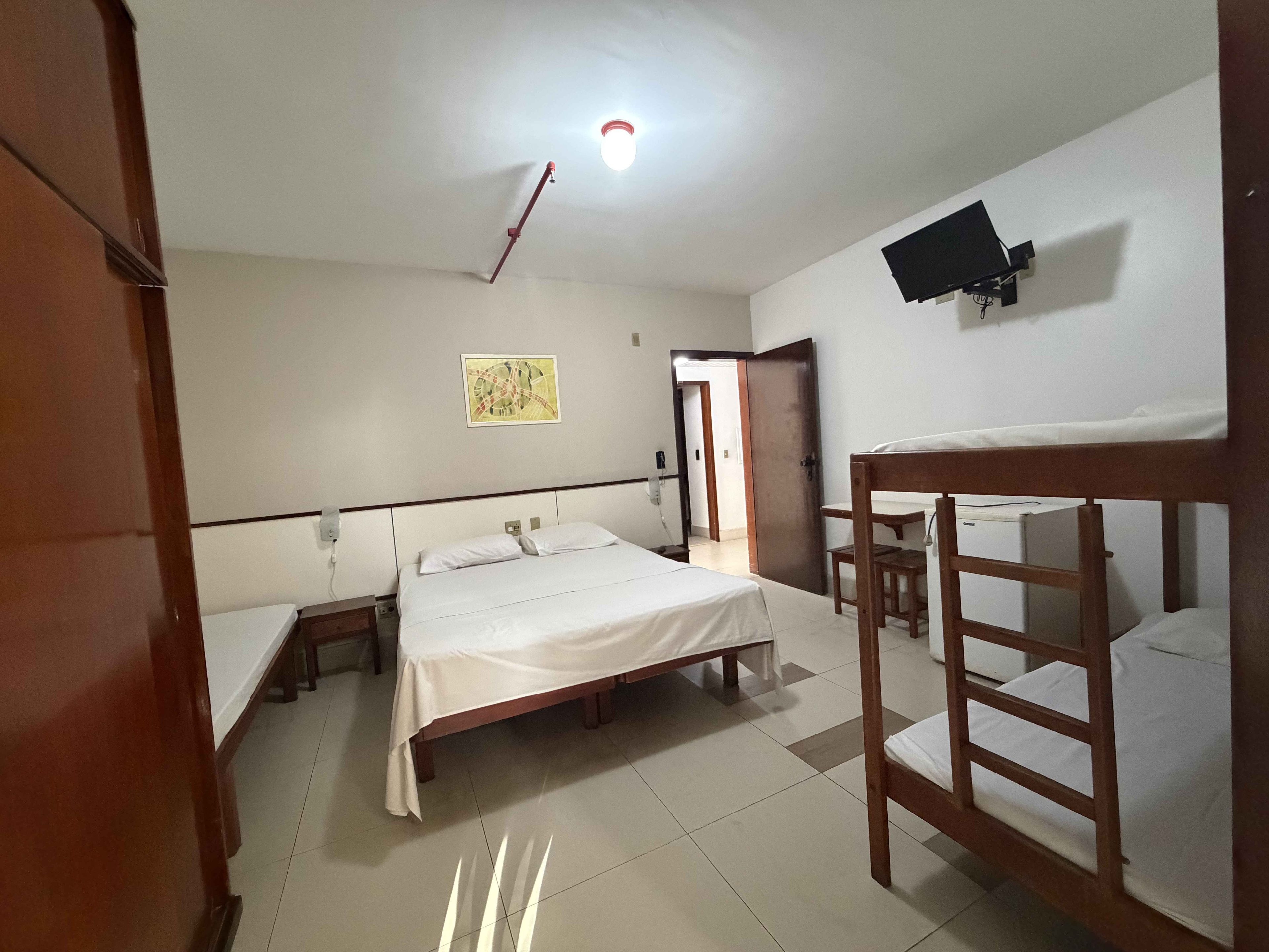 A room with a bed, a desk, a chair, a TV mounted on the wall, and a door leading to another room.