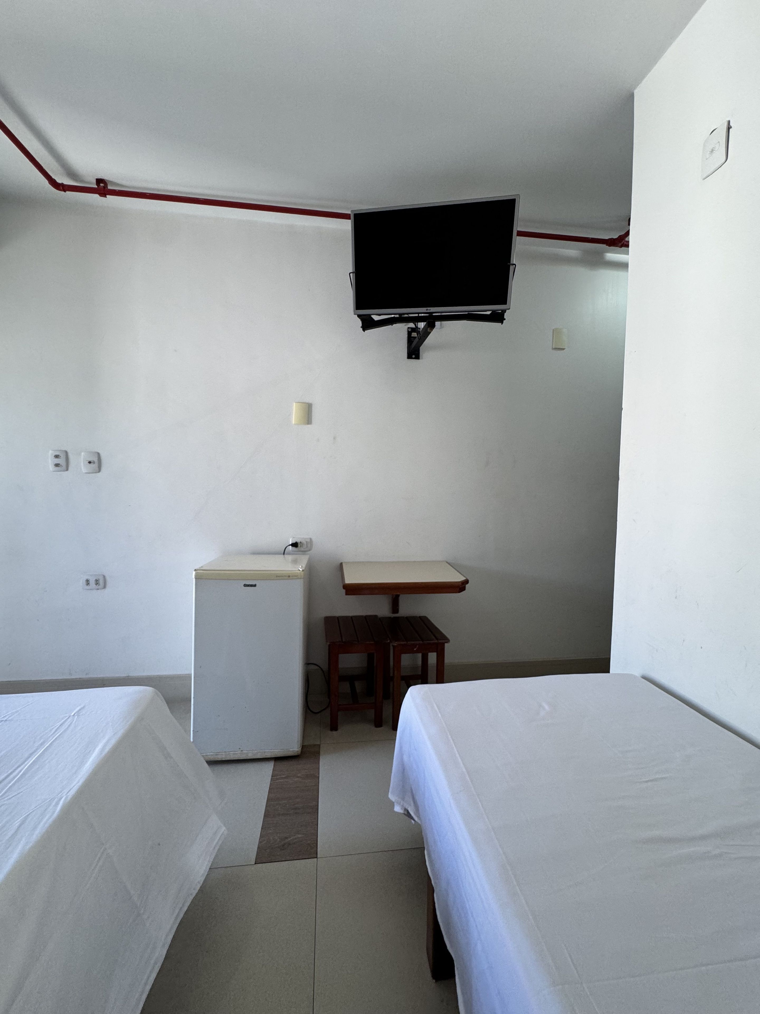 Una habitación pequeña y sencilla con dos camas, una mesa pequeña, un refrigerador y un televisor montado.
