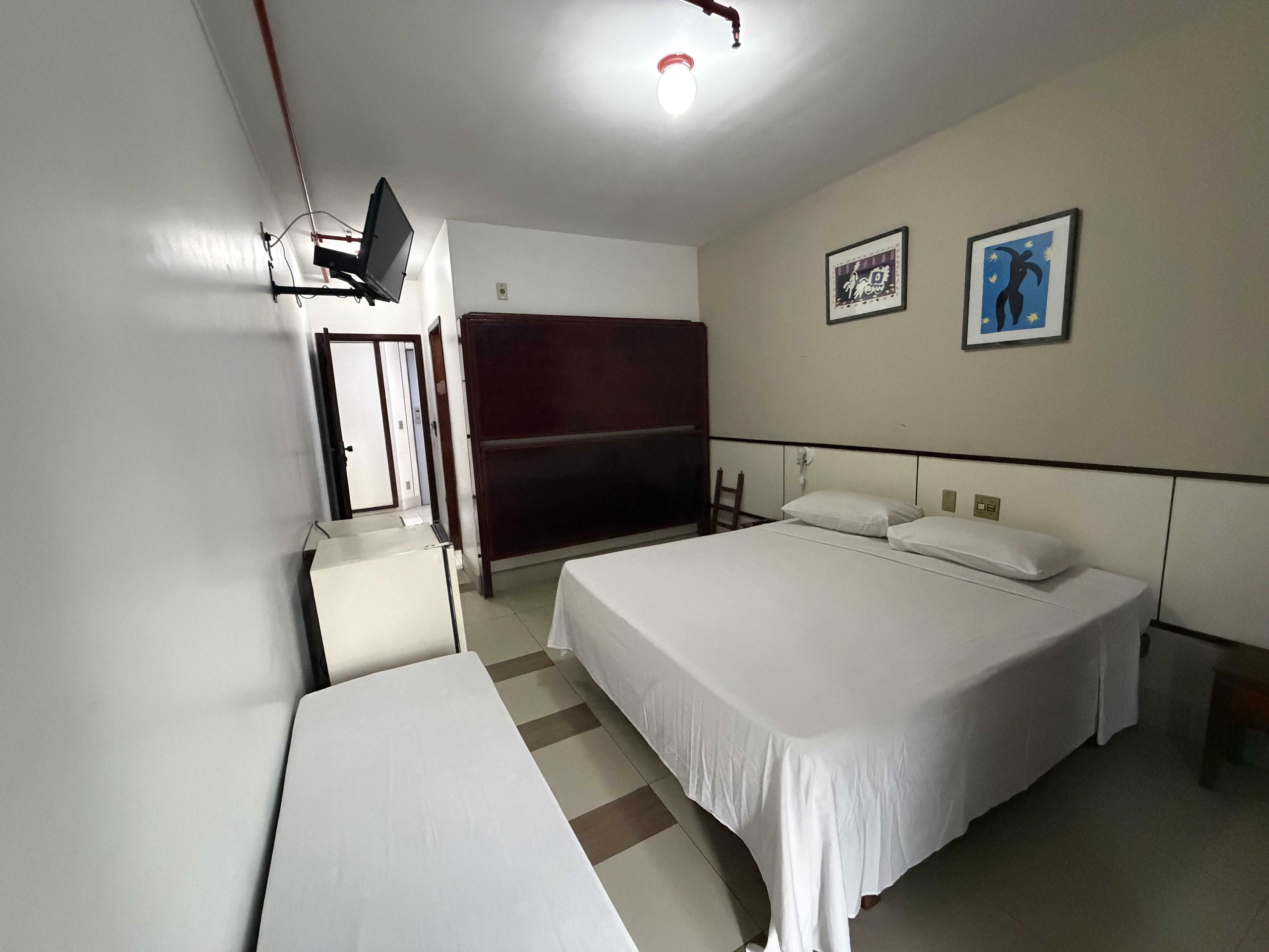 A hotel room with two beds, a desk, a chair, a wardrobe, a television, and two framed pictures on the wall.
