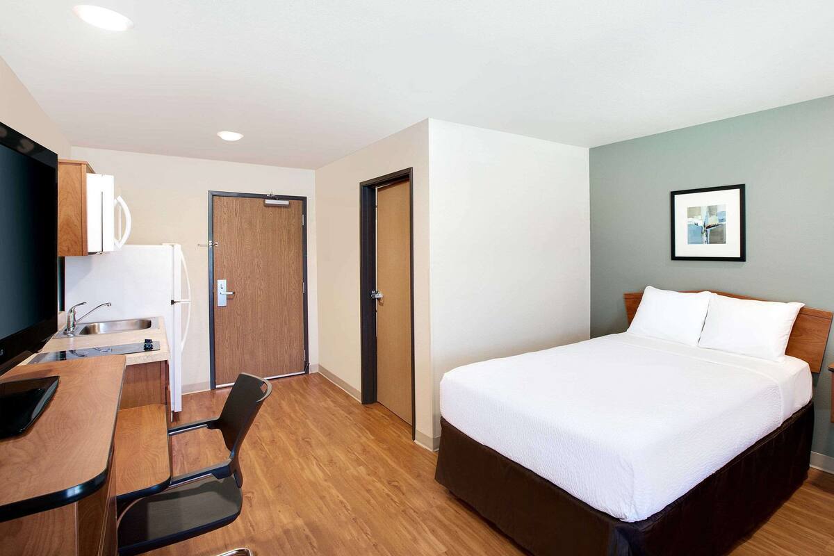 A hotel room with a bed, a desk with a chair, a kitchenette, and a door.