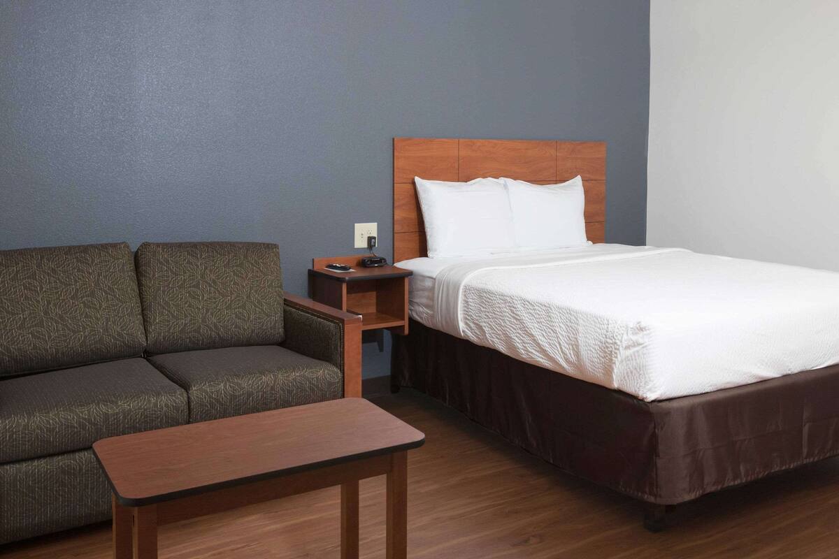 A hotel room with a bed, a sofa, a small wooden table, and a nightstand with a phone and a lamp.