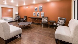 A modern living room with a brown wooden floor, beige sofas, a small round table, and a coffee table. There are four framed pictures on the orange wall. - WoodSpring Suites Lafayette (Lafayette)