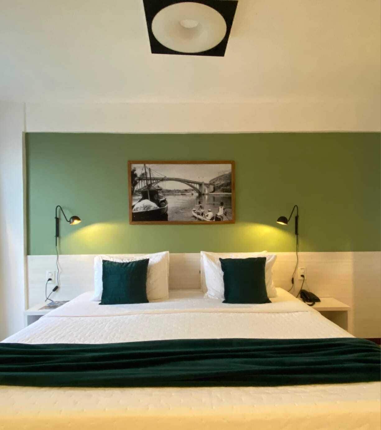 A hotel room with a bed, two bedside lamps, a nightstand, and a wall-mounted picture of a bridge.