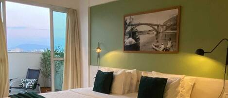 A bedroom with a large bed, a green wall, a painting of a bridge, and a balcony with a chair and plant.