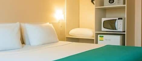 A hotel room with a bed, a green throw blanket, a white pillow, a wall-mounted lamp, a shelf with a microwave, and a framed picture on the wall.