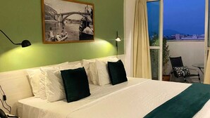 Une chambre d’hôtel avec un grand lit, une table de chevet, un bureau, une chaise, une fenêtre donnant sur la ville et un tableau au mur.