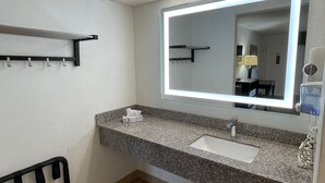 A modern bathroom with a granite countertop, a white sink, a large mirror with LED lighting, a towel rack, and a wooden floor. - Tricove Inn & Suites (Jacksonville)