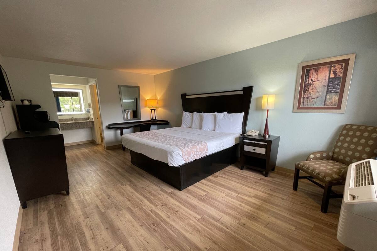 A hotel room with a large bed, a desk, a chair, and a small table with a lamp.