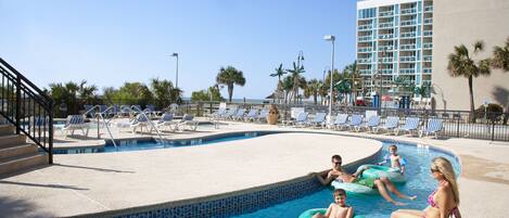 A pool area with lounge chairs, a building in the background, and a clear sky.