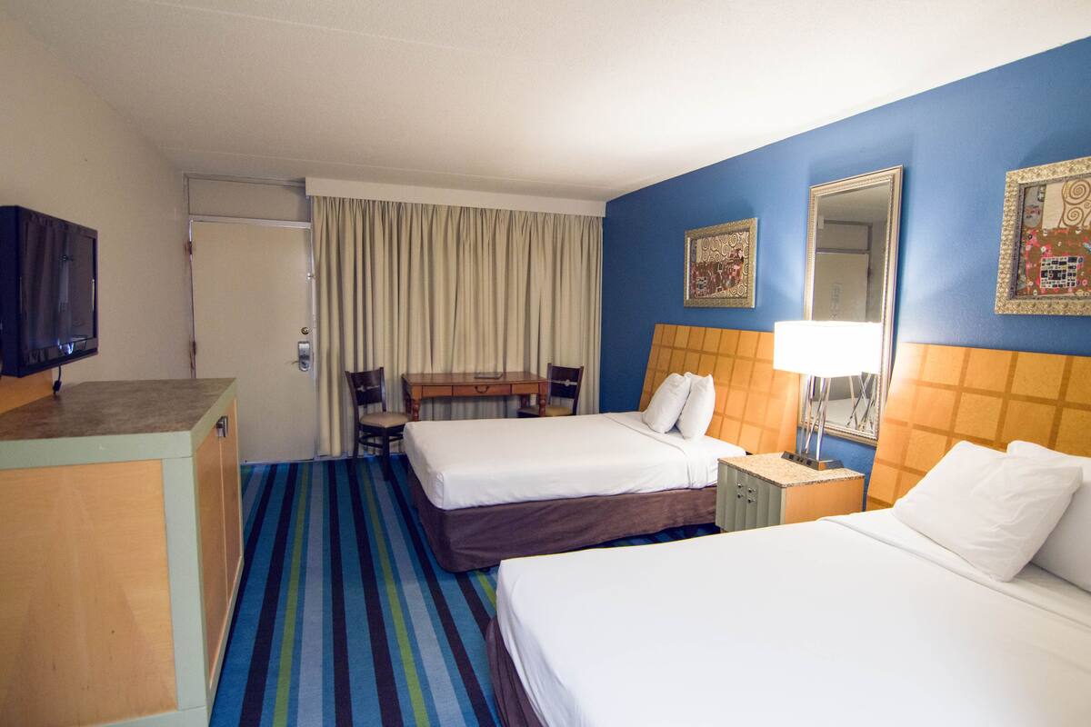 Une chambre d’hôtel avec deux lits, un bureau, une chaise, une télévision et une salle de bain équipée d’un miroir et de rideaux.