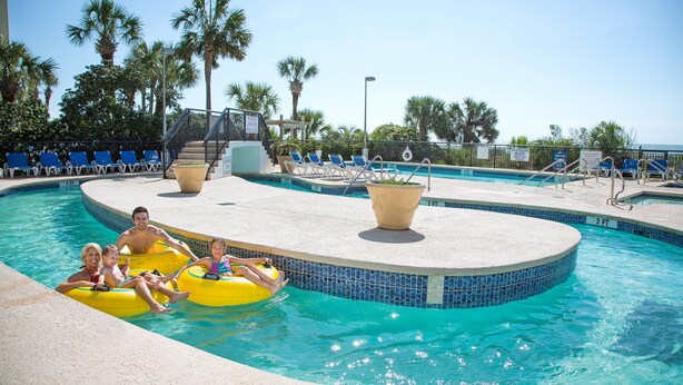 Un área de piscina con fuente circular, sillones reclinables y palmeras.