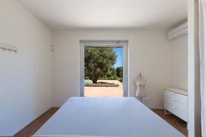 Ein Schlafzimmer mit einem großen Bett, einer Kommode und Blick auf einen Baum und Garten durch eine offene Tür.